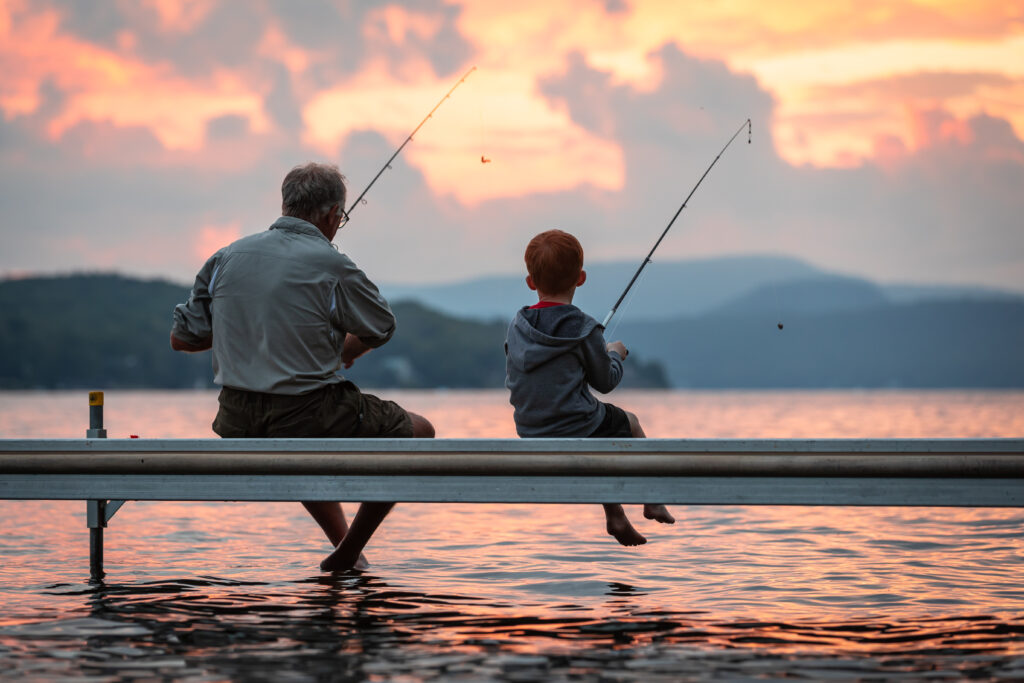 A grandfather is teaching his grandson to fish during summer. They are both sitting on the dock. It is a beautiful summer day at sunset. Across the lake, there is a mountain, Quebec, Canada.